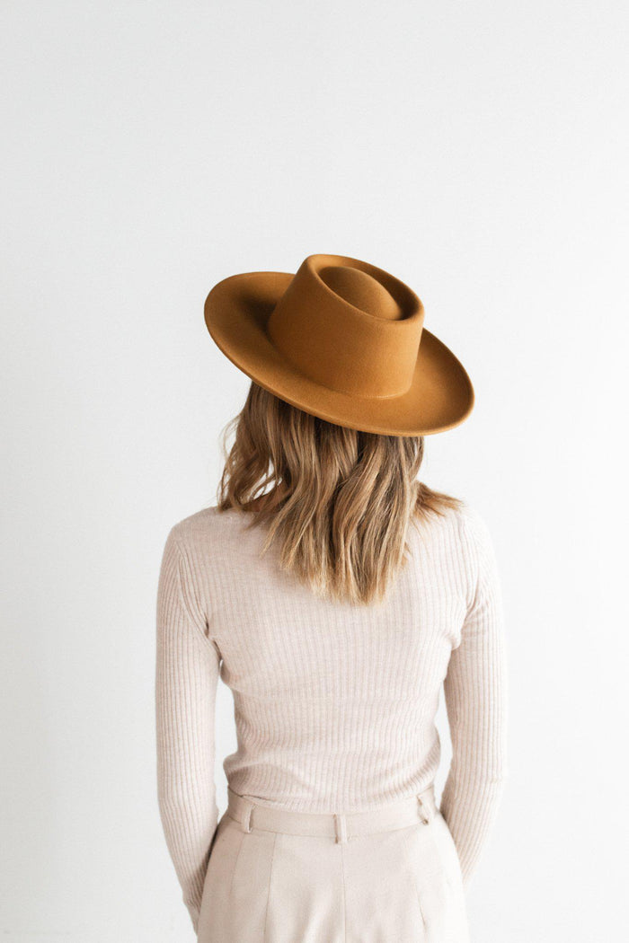 Woman wearing a cinnamon colored fedora hat, a cream top and white pants, facing behind against a plain background #color_cinnamon