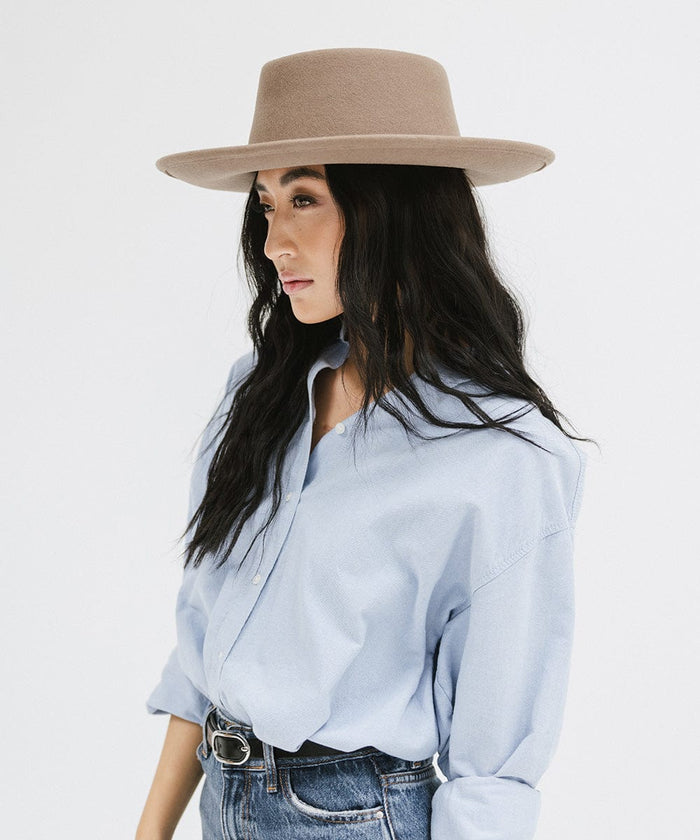 Woman wearing a taupe colored gambler hat, a blue shirt and jeans, facing sideways against a plain background #color_taupe