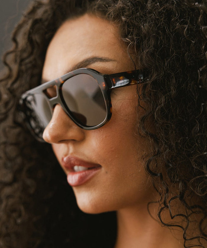 Woman with curly hair wearing tortoise colored aviator sunglasses against a dark grey background #color_tortoise