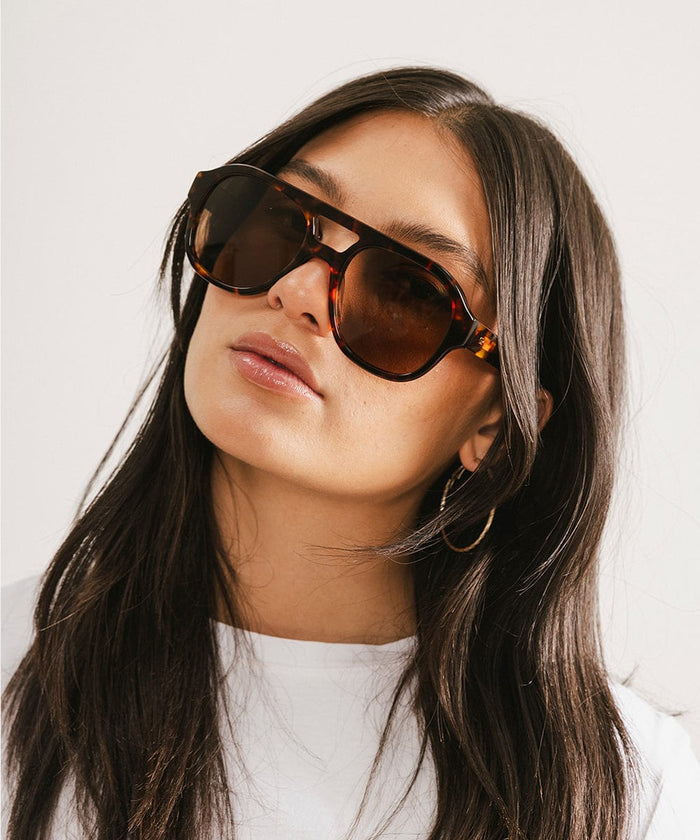 Woman wearing tortoise colored aviator sunglasses and a white t-shirt against a plain background #color_tortoise