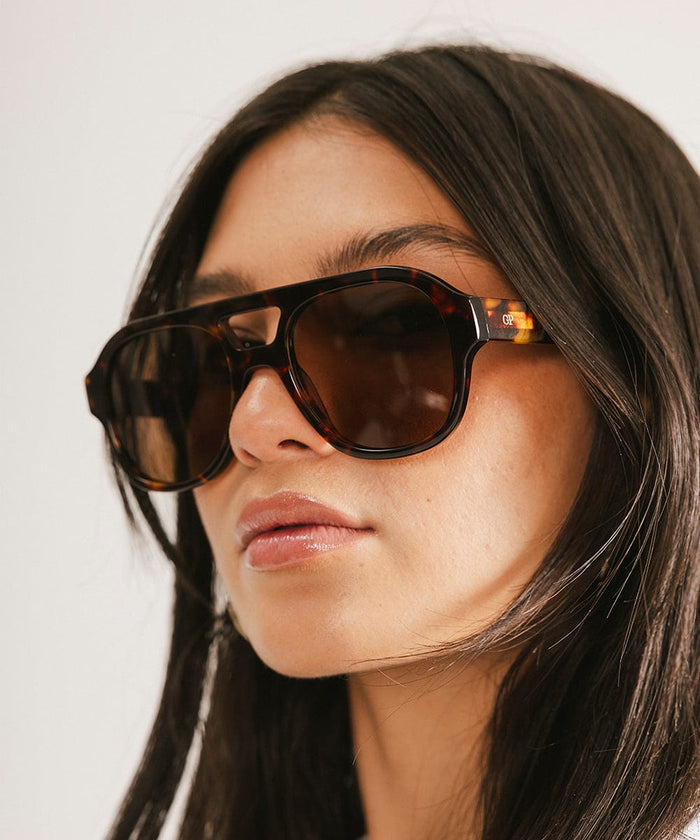Woman wearing tortoise colored aviator sunglasses against a plain  background #color_tortoise