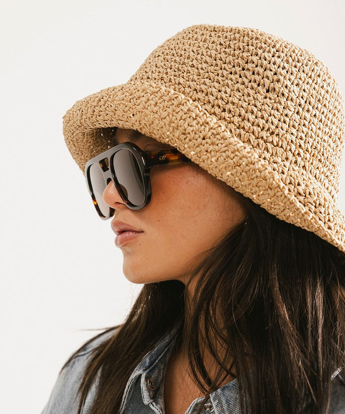 Woman wearing a brown hat and  tortoise colored aviator sunglasses, facing sideways against a plain background #color_tortoise