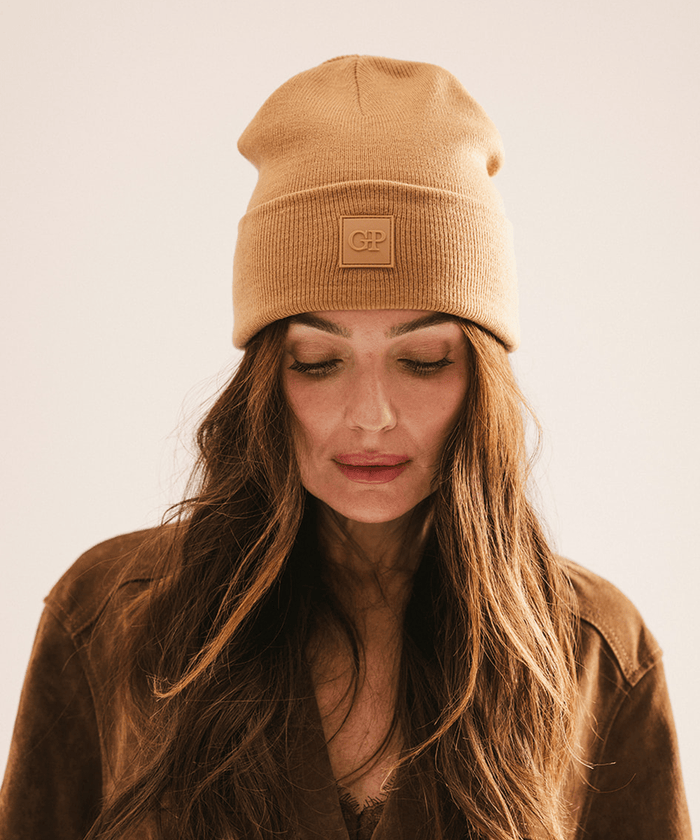 Woman wearing a latte colored beanie with a brand logo, looking down against a plain background #color_latte