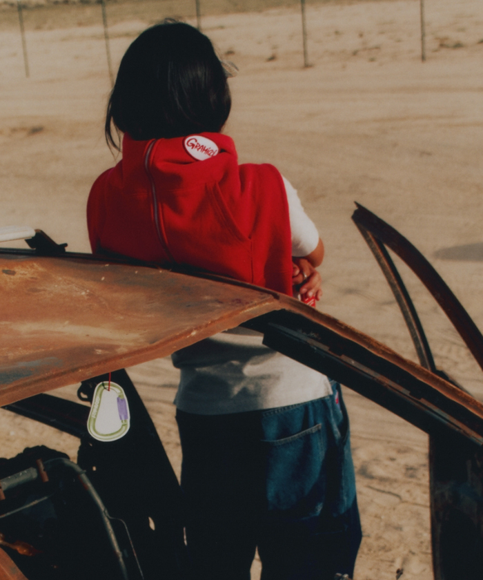 Model wearing a red Gramicci jacket eaning against an old car with an Olive Gramicci & retaW Carabiner Car Tag air freshener hanging from the rear-view mirror.