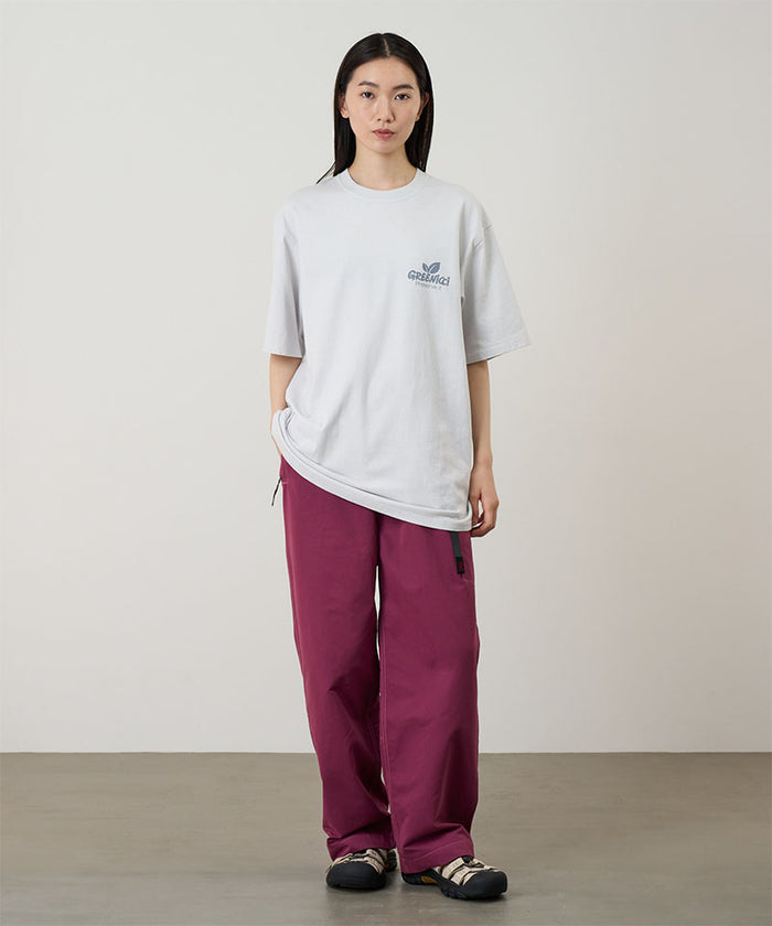Model wearing a smoke colored Gramicci Greenicci Tee with a graphic and purple pants on a light tone background.