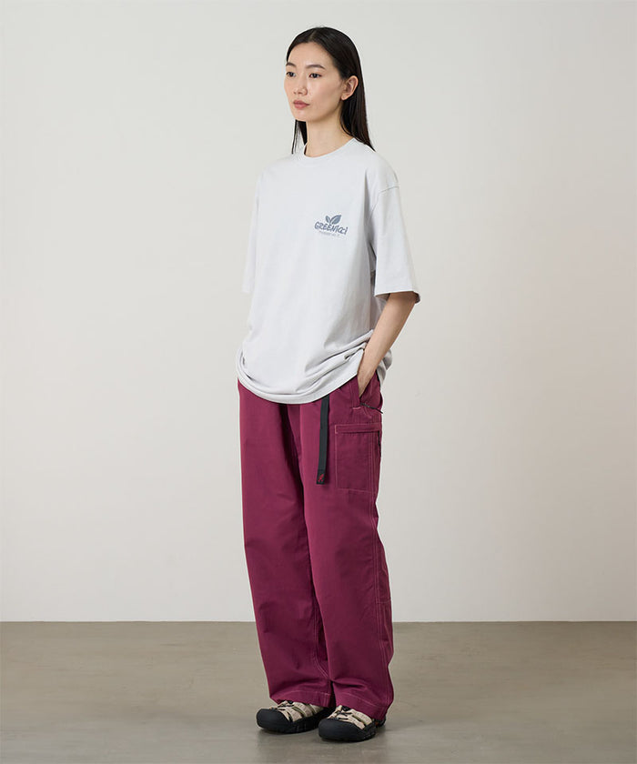Model wearing a smoke colored Gramicci Greenicci Tee with a graphic and purple pants on a light tone background.