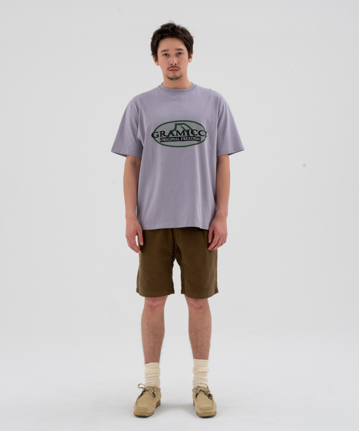 Model wearing a lavender Gramicci Half Dome Tee with green graphic and brown shorts on a light-tone background.