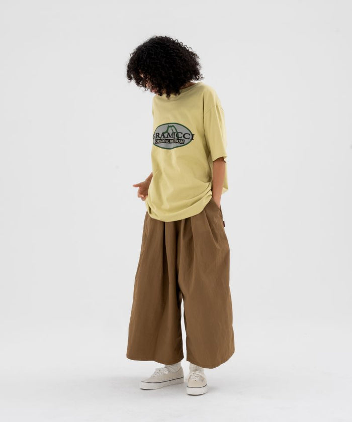 Model wearing a foggy lime Gramicci Half Dome Tee with grey graphic and brown pants on a light-tone background.