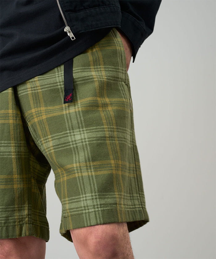 Model wearing olive plaid Gramicci Hemp G-Short Plaid and a black jacket on a light-tone background.