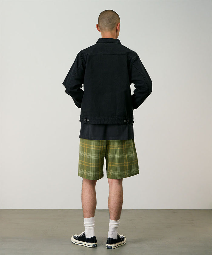 Back view of a model wearing olive plaid Gramicci Hemp G-Short Plaid and a black jacket on a light-tone background.