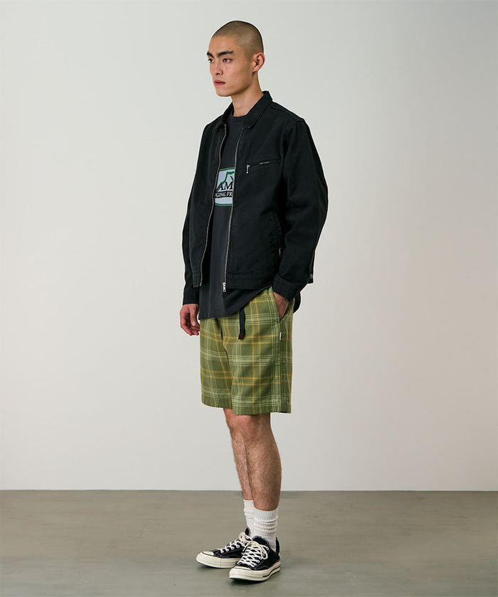 Model wearing olive plaid Gramicci Hemp G-Short Plaid and a black jacket on a light-tone background.