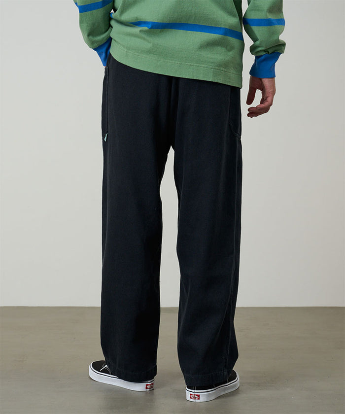 Back view of a model wearing carbon colored Gramicci Hemp Relaxed Pants and a green and blue striped shirt on a light-tone background.
