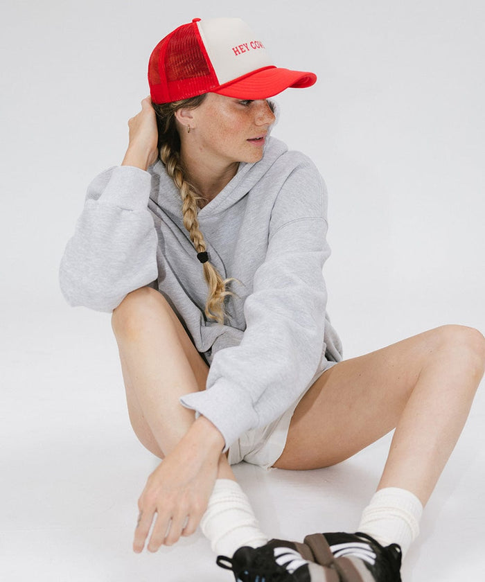 Woman wearing a cream and vintage red cap with 'Hey Cowboy' text and a grey sweater, sitting against a plain background  #color_cream - vintage red