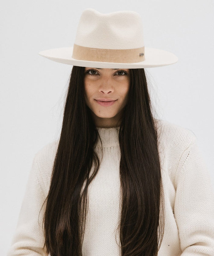 Woman with long hair wearing an off white felt fedora hat with a band and a white sweater against a plain background  #color_off white