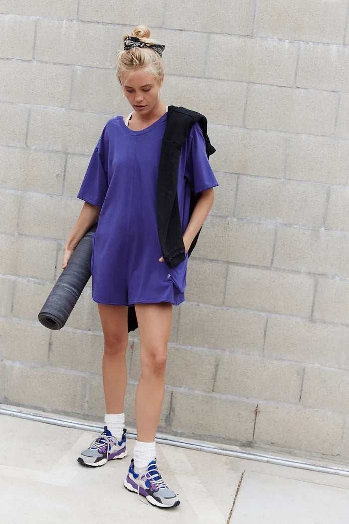 Woman standing in Free People Hot Shot Tee Romper Coddy in the color Dark Amethyst holding rolled yoga mat
