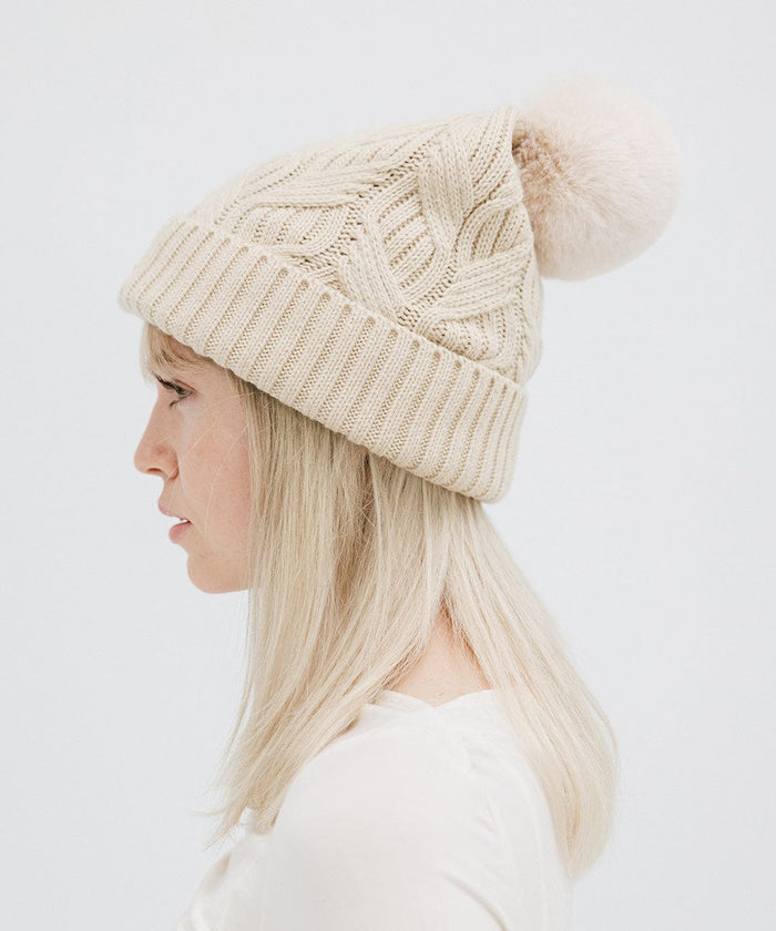 Woman wearing a cream knit beanie with a pom-pom and a white top, facing sideways against a plain background #color_cream