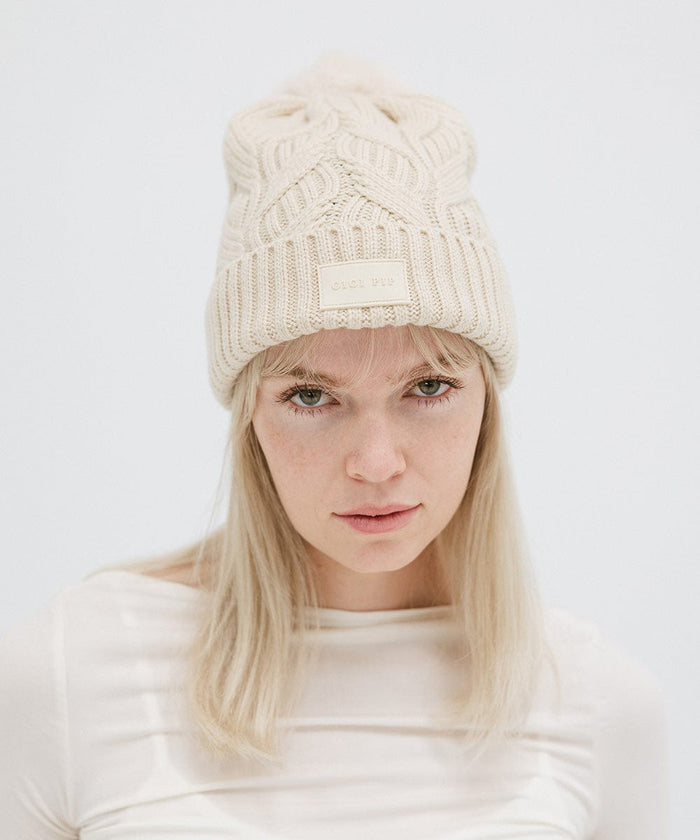 Woman wearing a cream knit beanie with a pom-pom and a white top against a plain background #color_cream