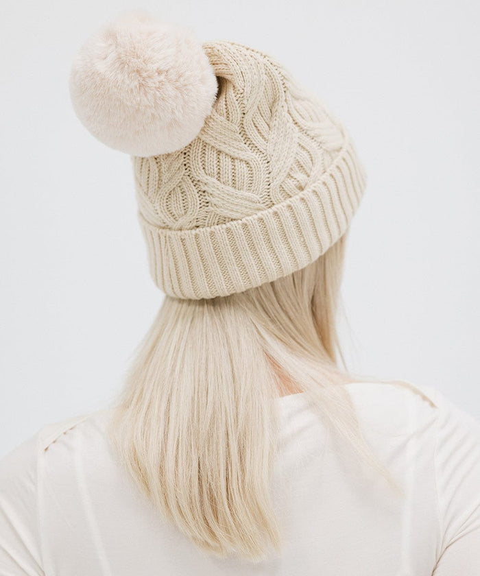 Woman wearing a cream knit beanie with a pom-pom and a white top, facing behind against a plain background #color_cream