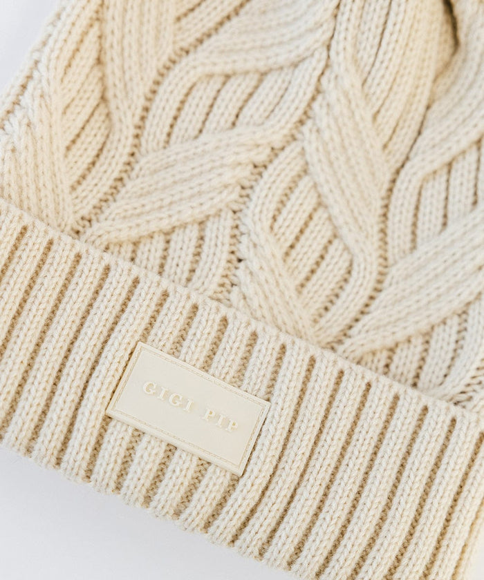 Close up image of a cream knit beanie with with the brand label on a plain background #color_cream