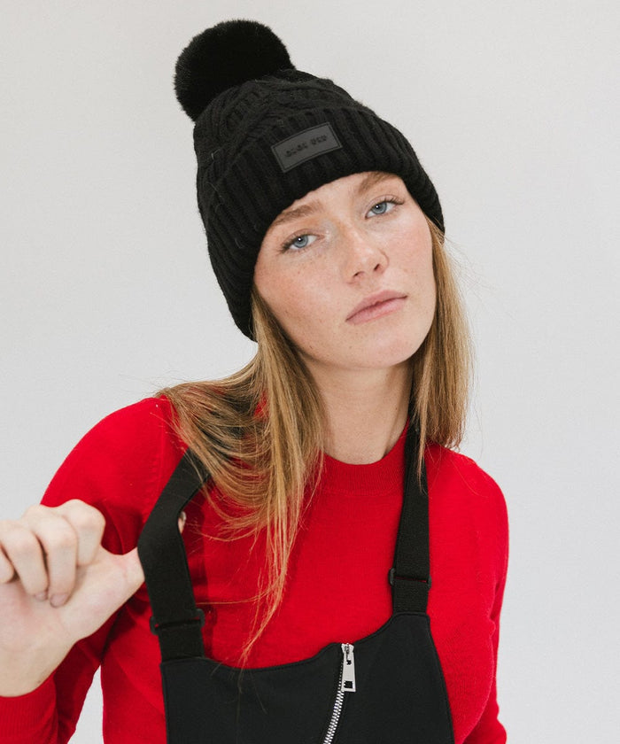Woman wearing a black knit beanie with a pom-pom and a red t-shirt with a black sleeveless jacket against a plain background #color_black