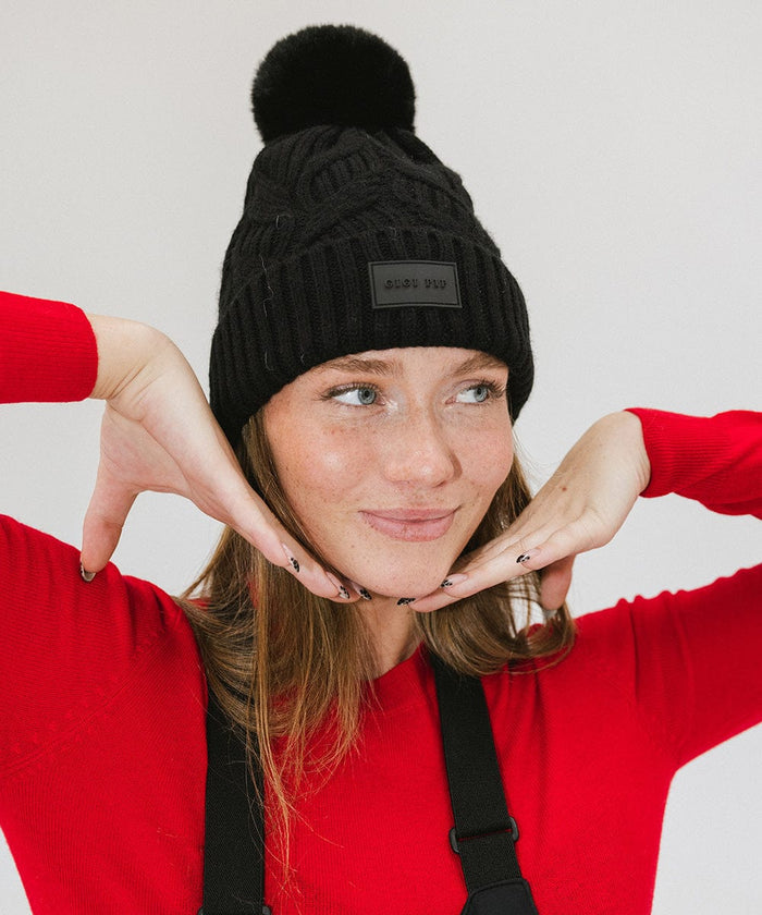 Woman wearing a black knit beanie with a pom-pom and a red t-shirt, posing against a plain background #color_black