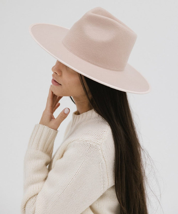 Woman wearing a pale nude colored fedora hat and a white sweater, posing sideways against a plain background #color_pale nude