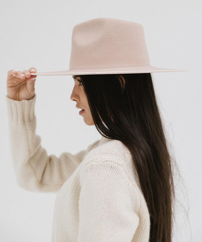 Woman wearing a pale nude colored fedora hat and a white sweater, facing sideways against a plain background #color_pale nude