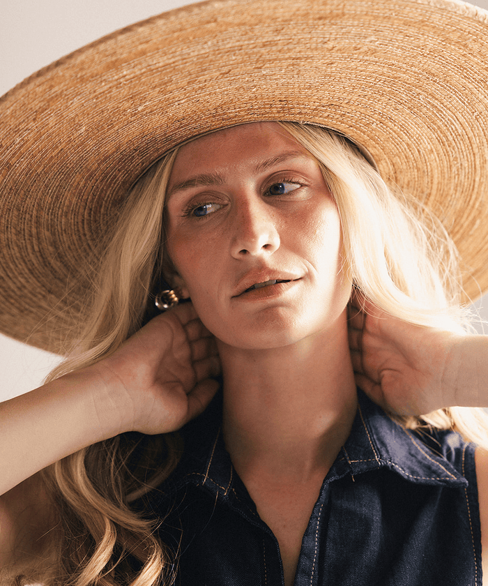 Woman wearing a wide brim boater straw hat against a white background #color_natural brown