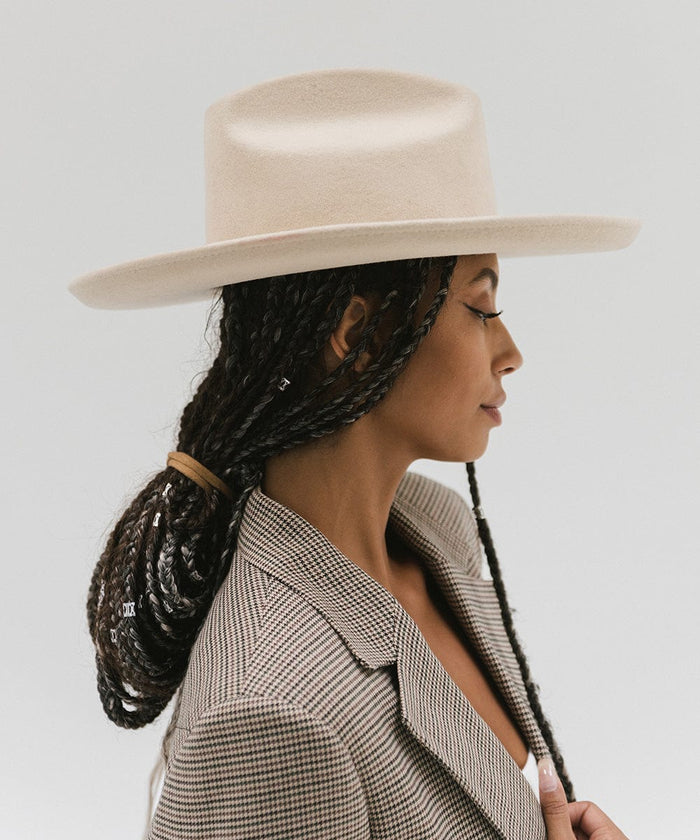 Woman wearing an off white western hat with a pencil brim and a tall crown and a coat, facing sideways against a plain background #color_off white