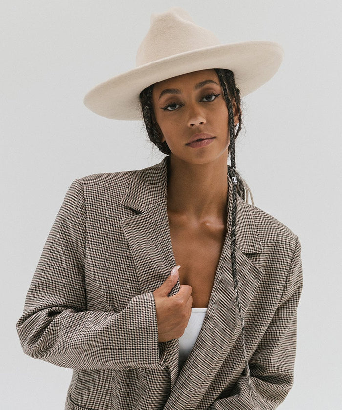 Woman wearing an off white western hat with a pencil brim and a tall crown and a coat against a plain background #color_off white