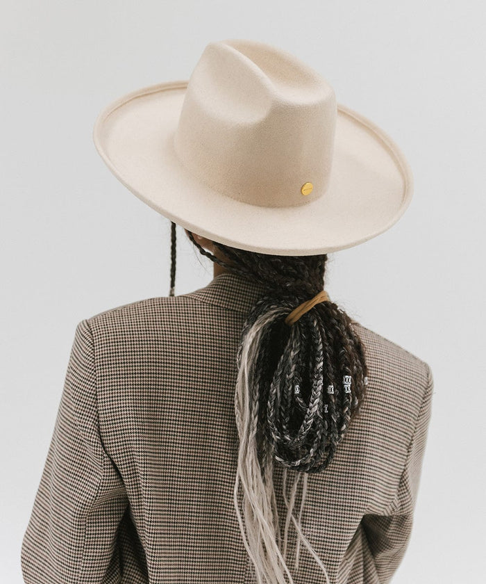 Person wearing an off white western hat with a pencil brim and a tall crown and coat against a plain background #color_off white