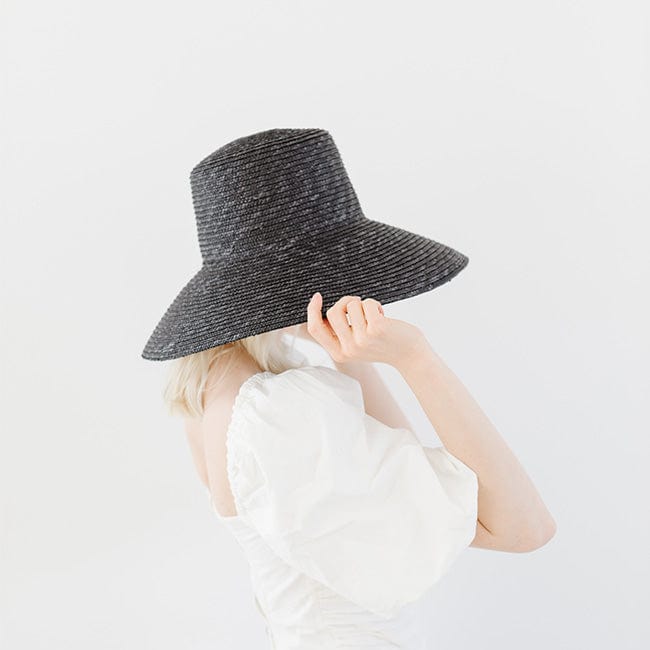 Woman wearing a black straw boater hat and a white top, facing sideways  against a plain background #color_black