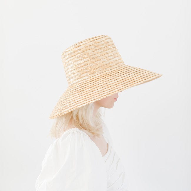 Woman wearing a natural colored straw boater hat and a white top, facing sideways against a plain background  #color_natural
