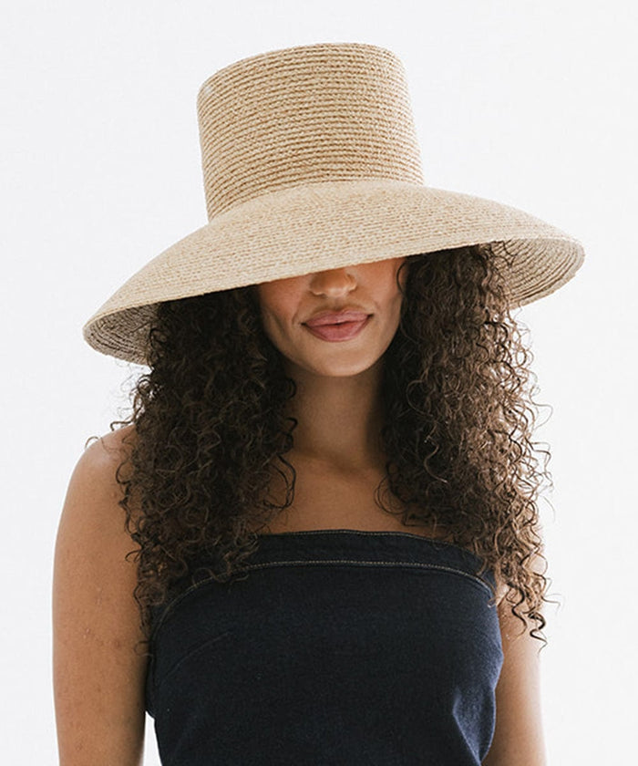 Woman with curly hair wearing a natural colored oversized packable straw bucket hat and a denim top against a plain background #color_natural