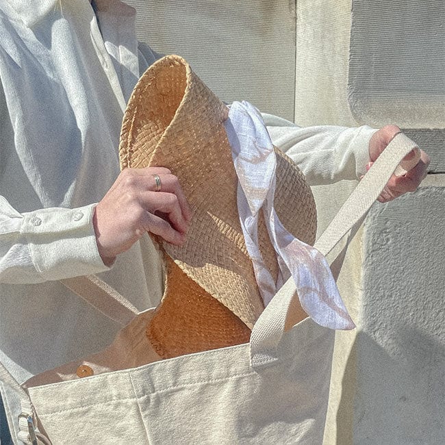 Person wearing a white shirt putting a natural colored packable straw hat inside a white bag against a white wall in the background #color_natural