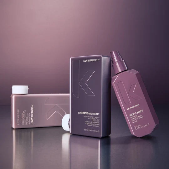 Kevin Murphy haircare products displayed on a reflective surface against a dark mauve background, including a Hydrate-Me Rinse bottle, a Night Shift spray bottle, and a matching box.