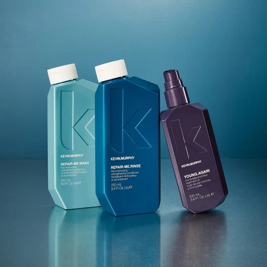 Three Kevin Murphy haircare bottles on a reflective blue surface against a blue gradient background: a light aqua Repair Me Wash bottle, a medium blue Repair Me Rinse bottle, and a dark purple Young Again spray bottle.