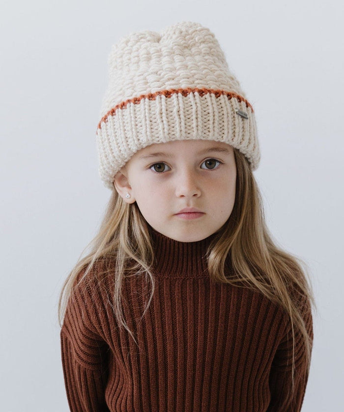 Child wearing a cream vail beanie with a burnt orange stripe and a brown sweater against a plain background #color_cream-burnt orange