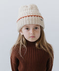 Child wearing a cream vail beanie with a burnt orange stripe and a brown sweater against a plain background #color_cream-burnt orange
