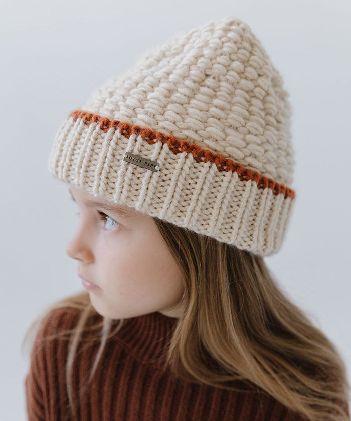 Child wearing a cream vail beanie with a burnt orange stripe and a brown sweater, looking sideways against a plain background #color_cream-burnt orange