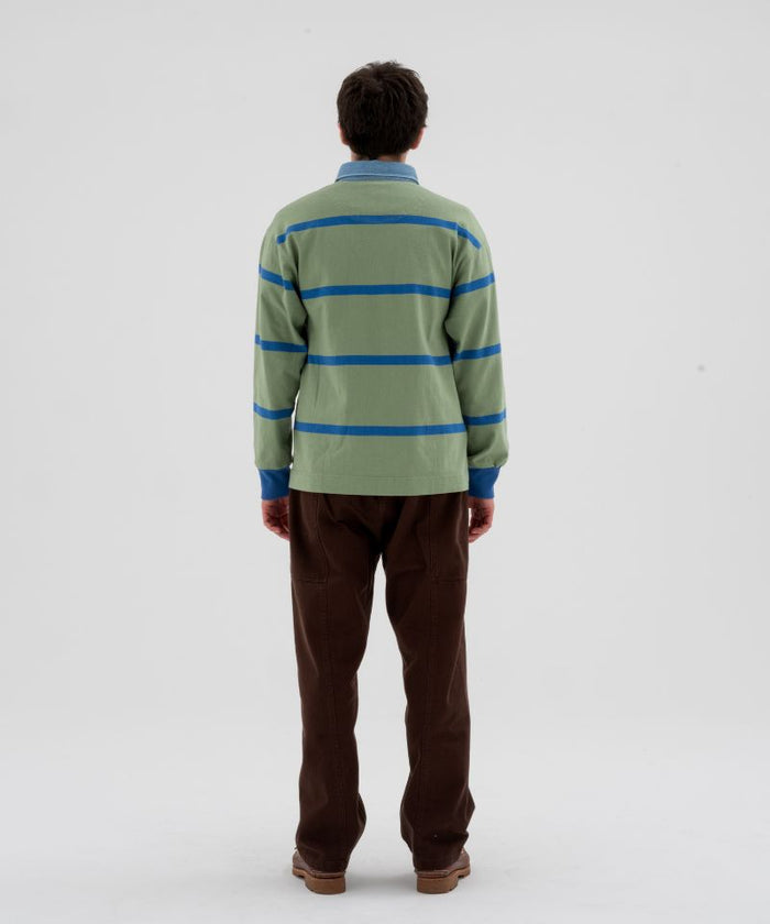 Back view of a model wearing a olive and blue striped Gramicci Langdon Rugby Shirt and brown pants on a light-tone background.