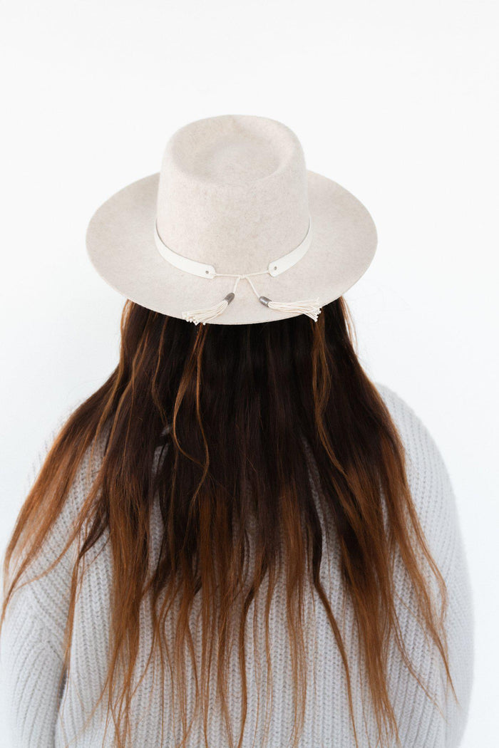 Woman wearing an ivory colored hat with an off white leather band with tassels and a white sweater, facing behind against a plain background #color_off white
