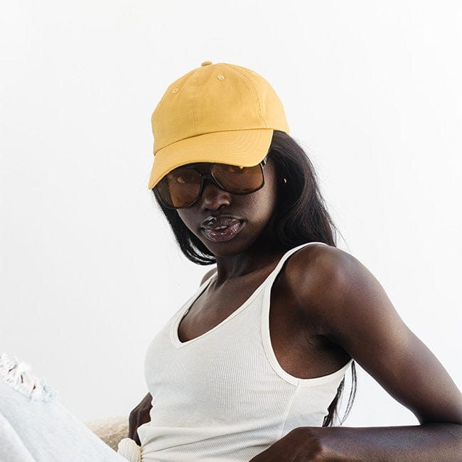 Woman wearing a mustard colored ball cap, sunglasses and a white top, posing against a plain background #color_mustard