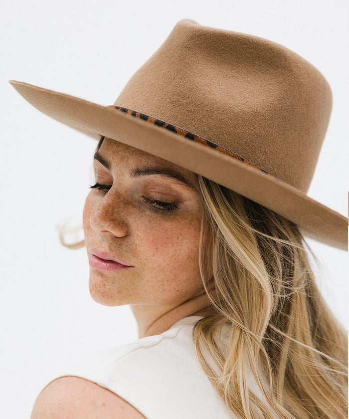 Woman wearing a beige hat with a dark brown leopard print hat band and a white top, posing sideways against a plain background #color_dark brown