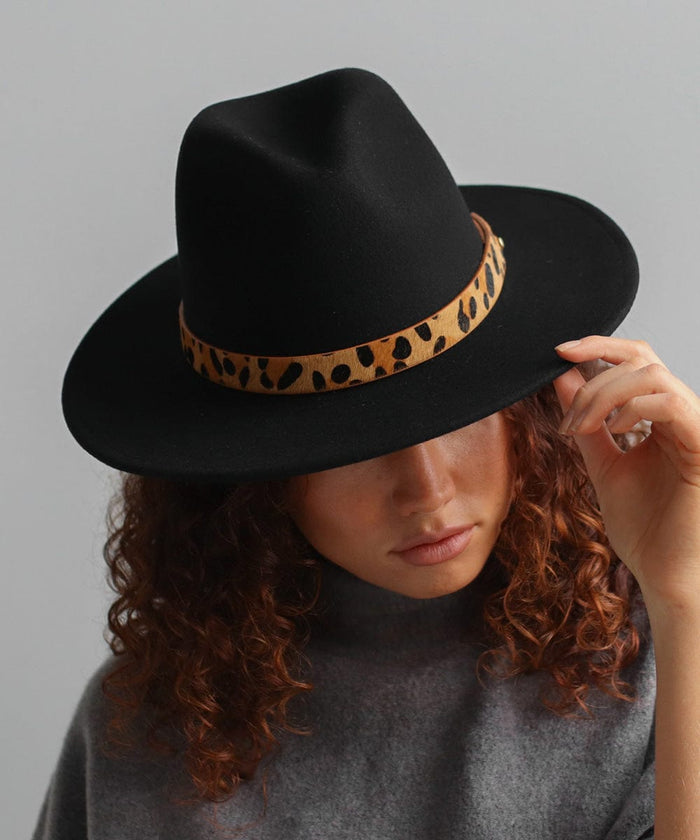 Woman wearing a black hat with a tan leopard print hat band and a grey t-shirt, looking down against a plain background #color_tan