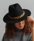 Woman wearing a black hat with a tan leopard print hat band and a grey t-shirt, looking down against a plain background #color_tan