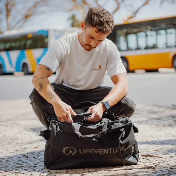 Man unpacking a Lifeventure Expedition Cargo Duffle Bag 50 Litres outdoors with buses in the background