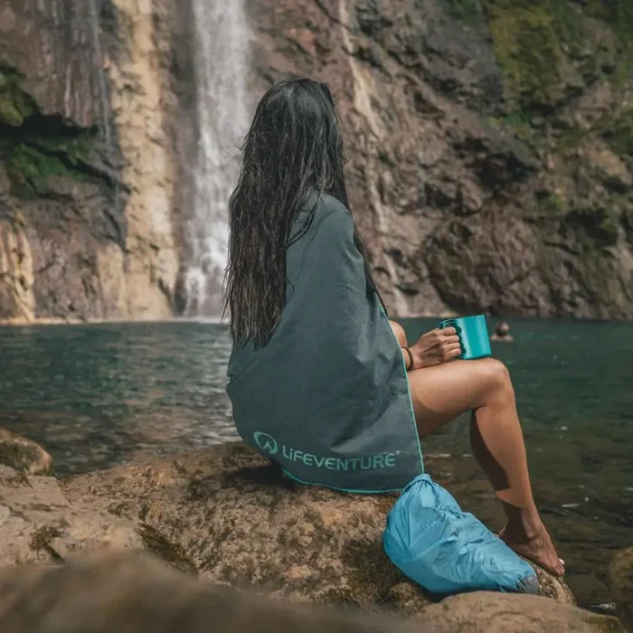 Woman sitting by a waterfall with a Lifeventure giant SoftFibre recycled travel towel wrapped around her shoulders and upper body