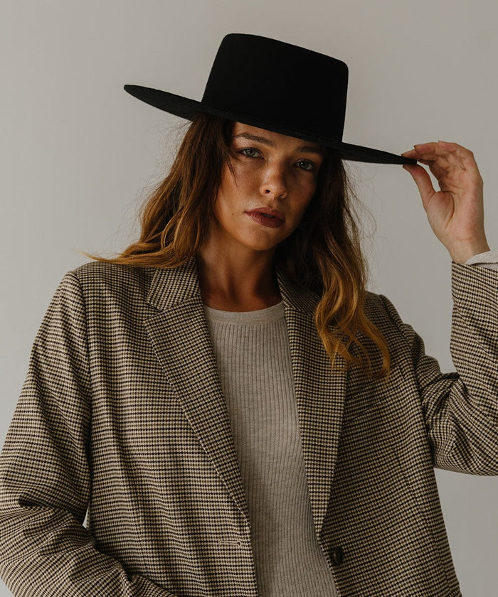 Woman wearing a black boater hat and a white top with a coat against a plain background #color_black
