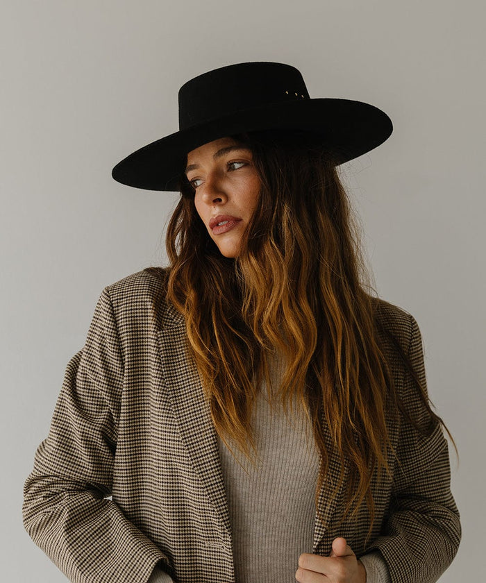 Woman wearing a black boater hat and a white top with a coat, facing sideways against a plain background #color_black